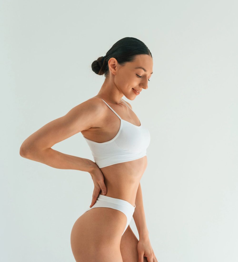 Relaxed, standing against white background. Young woman with slim body type is in fitness clothes in the studio.